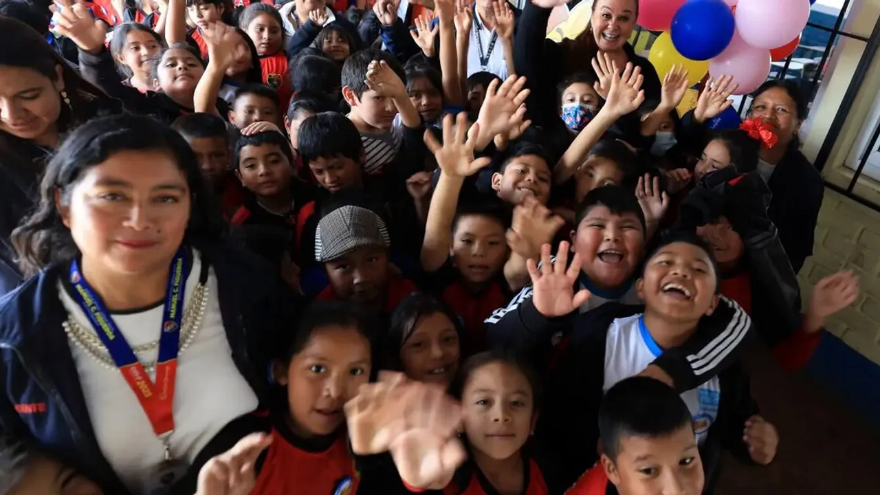 Docentes y niños sonrientes en un aula levantan las manos para celebrar el cierre del año escolar.