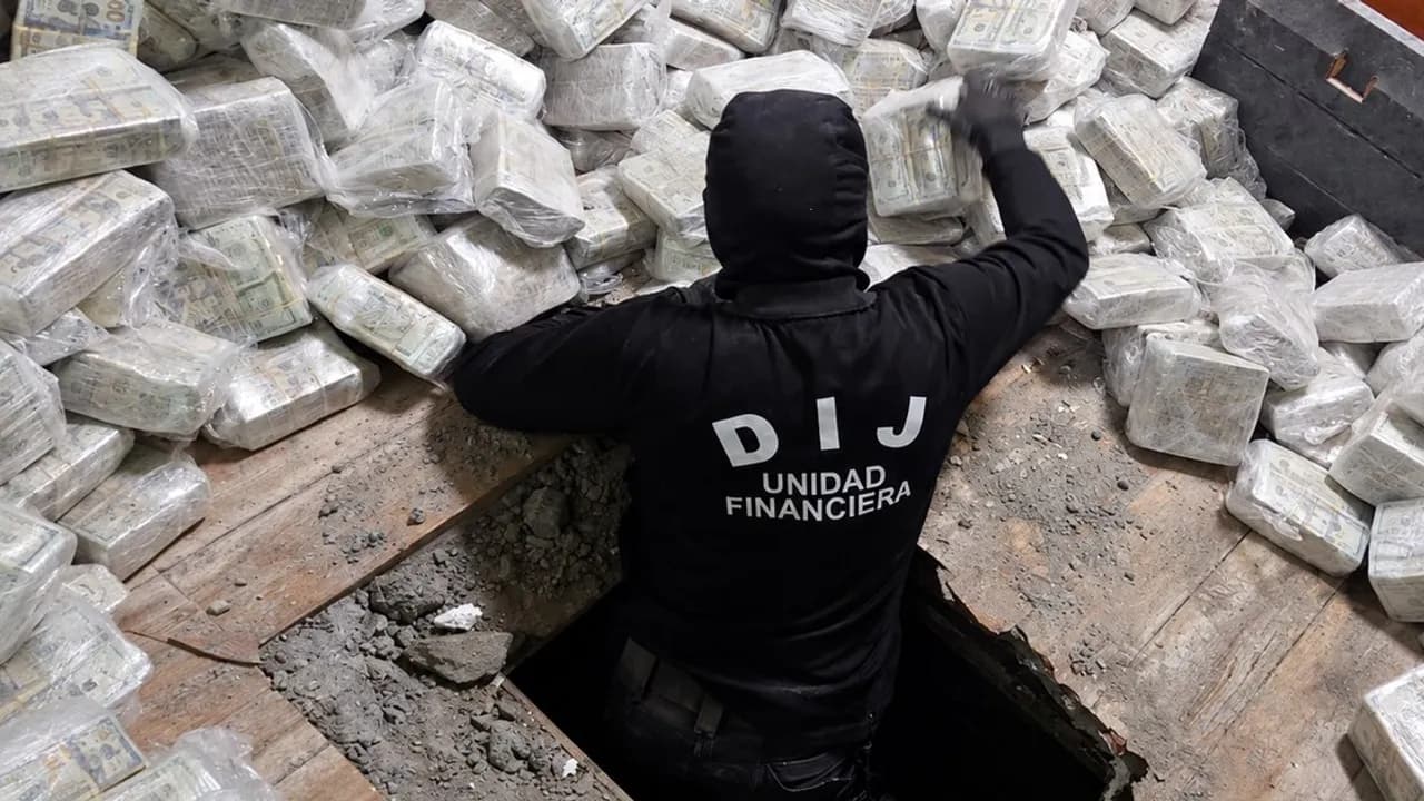 Agente policial de espalda junto a una caleta subterránea repleta de fajos de dólares incautados en paquetes plásticos.