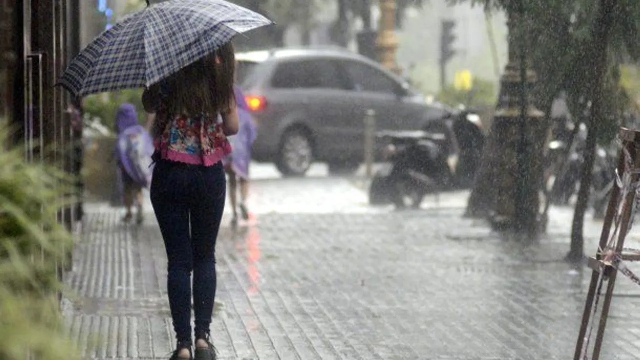 Persona con paraguas camina bajo una intensa lluvia sobre una vereda mojada mientras circula un automóvil.