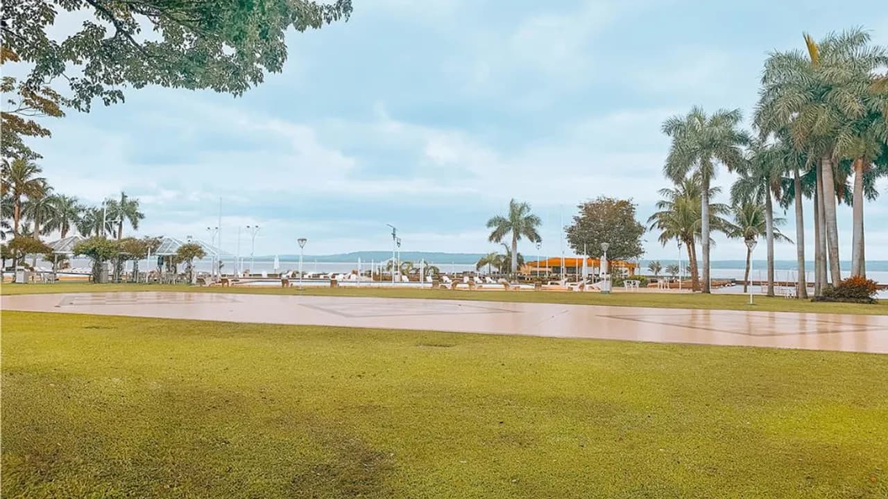 Parque con césped, palmeras y explanada junto al lago bajo un cielo nublado en las instalaciones ribereñas.