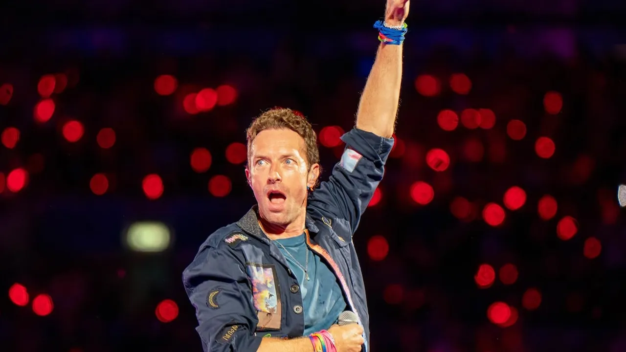 Chris Martin canta con el micrófono en mano y el brazo levantado ante un fondo iluminado de luces rojas.