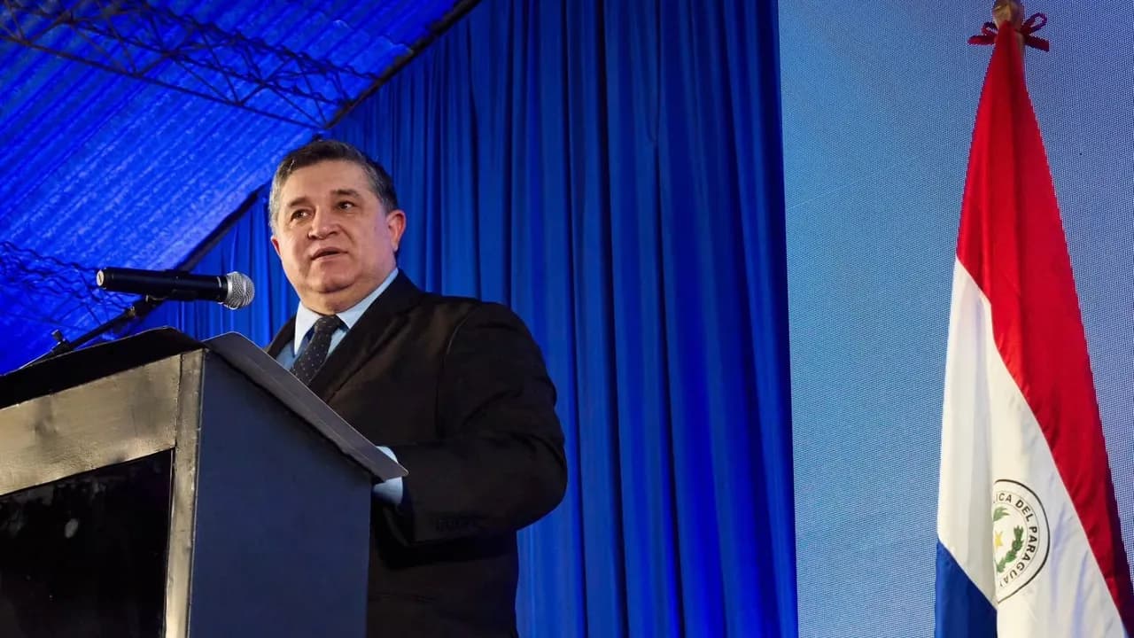 Hombre de traje oscuro habla desde un podio frente a un micrófono, con telón azul y una bandera paraguaya al costado.