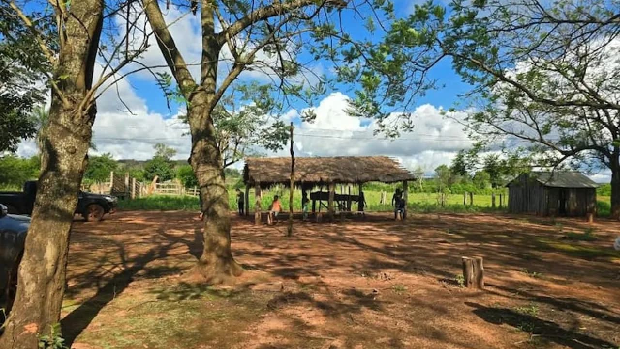 Patio de tierra con choza de paja y árboles en un asentamiento rural bajo cielo azul con nubes.