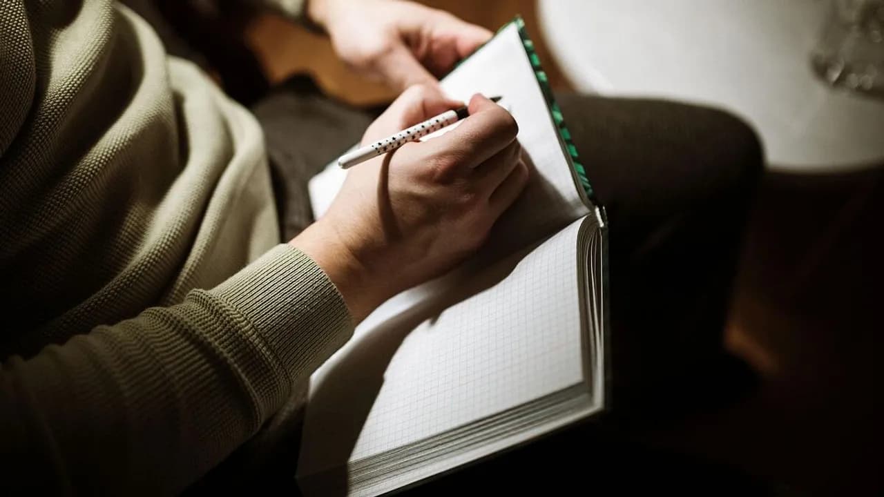 Manos de una persona sosteniendo un bolígrafo mientras redacta textos en una libreta de hojas cuadriculadas.