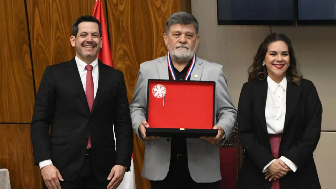 Ricardo Flecha sostiene un estuche rojo con una medalla, acompañado por dos autoridades en una sala oficial.
