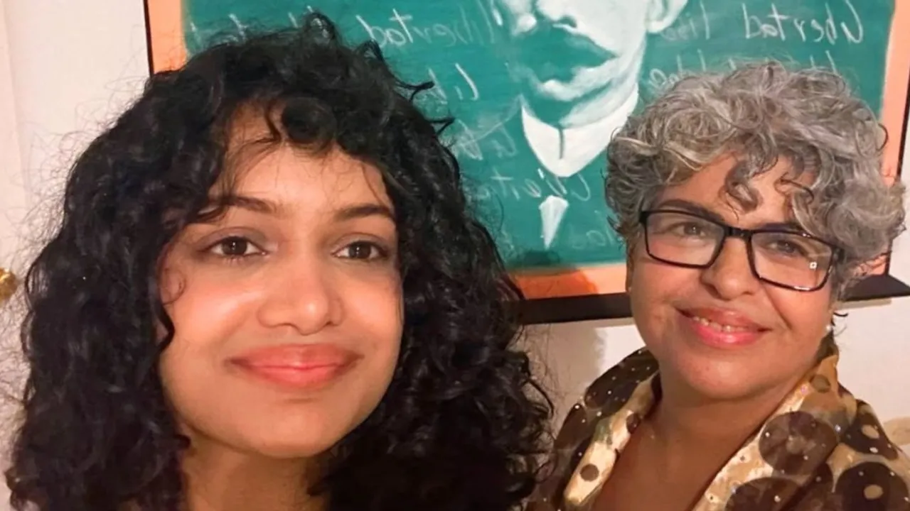 Dos mujeres sonrientes de cabello rizado posan juntas frente a una pintura verde de un rostro.