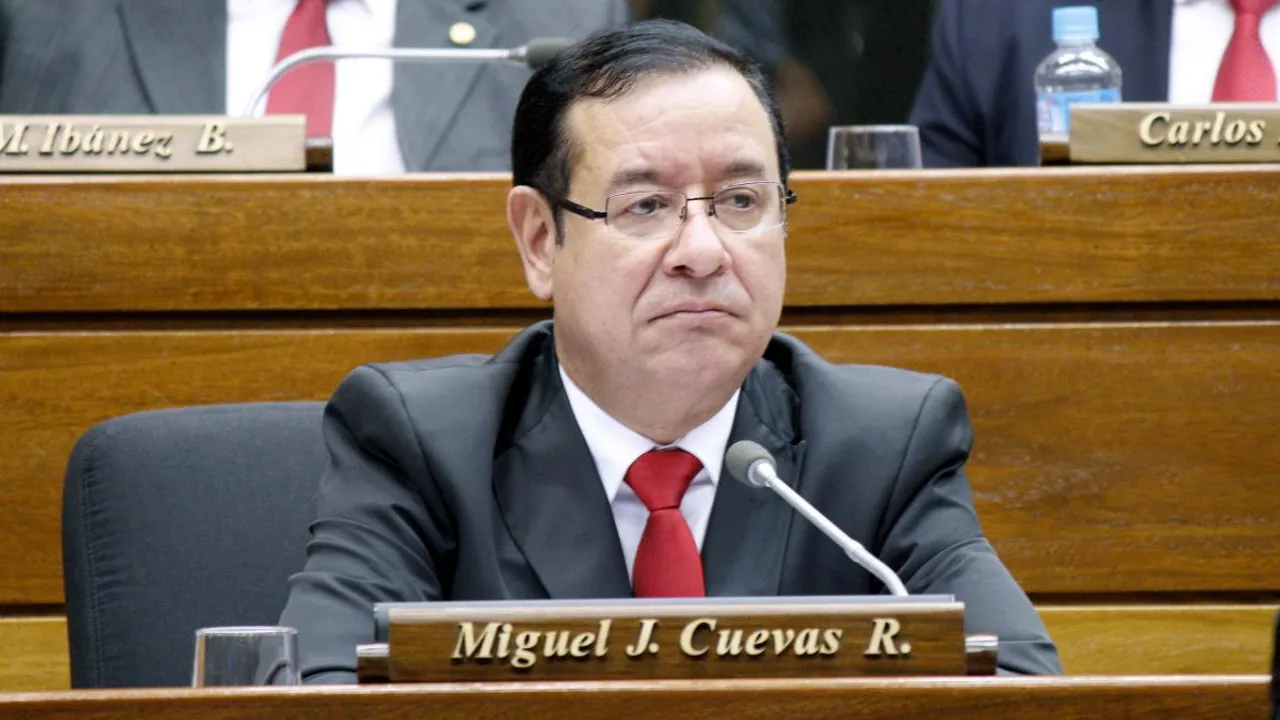 Miguel Cuevas con anteojos, traje oscuro y corbata roja en una banca legislativa con placa con su nombre.