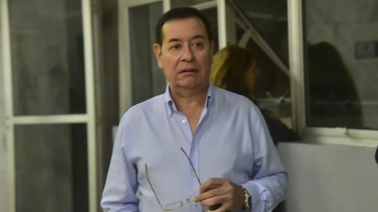 Hombre con camisa celeste de mangas largas sostiene sus anteojos en la mano dentro de una oficina iluminada.