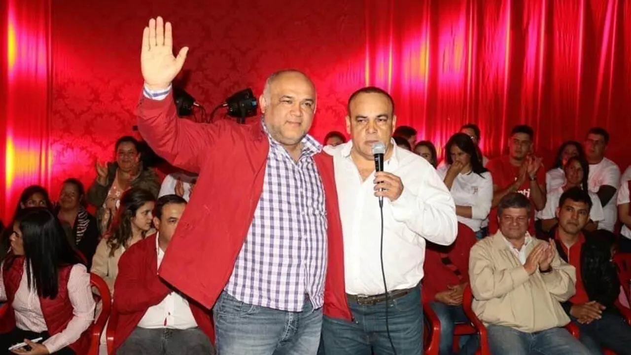 Dos hombres en un acto público frente a un telón rojo, uno de ellos con micrófono y el otro levantando la mano.