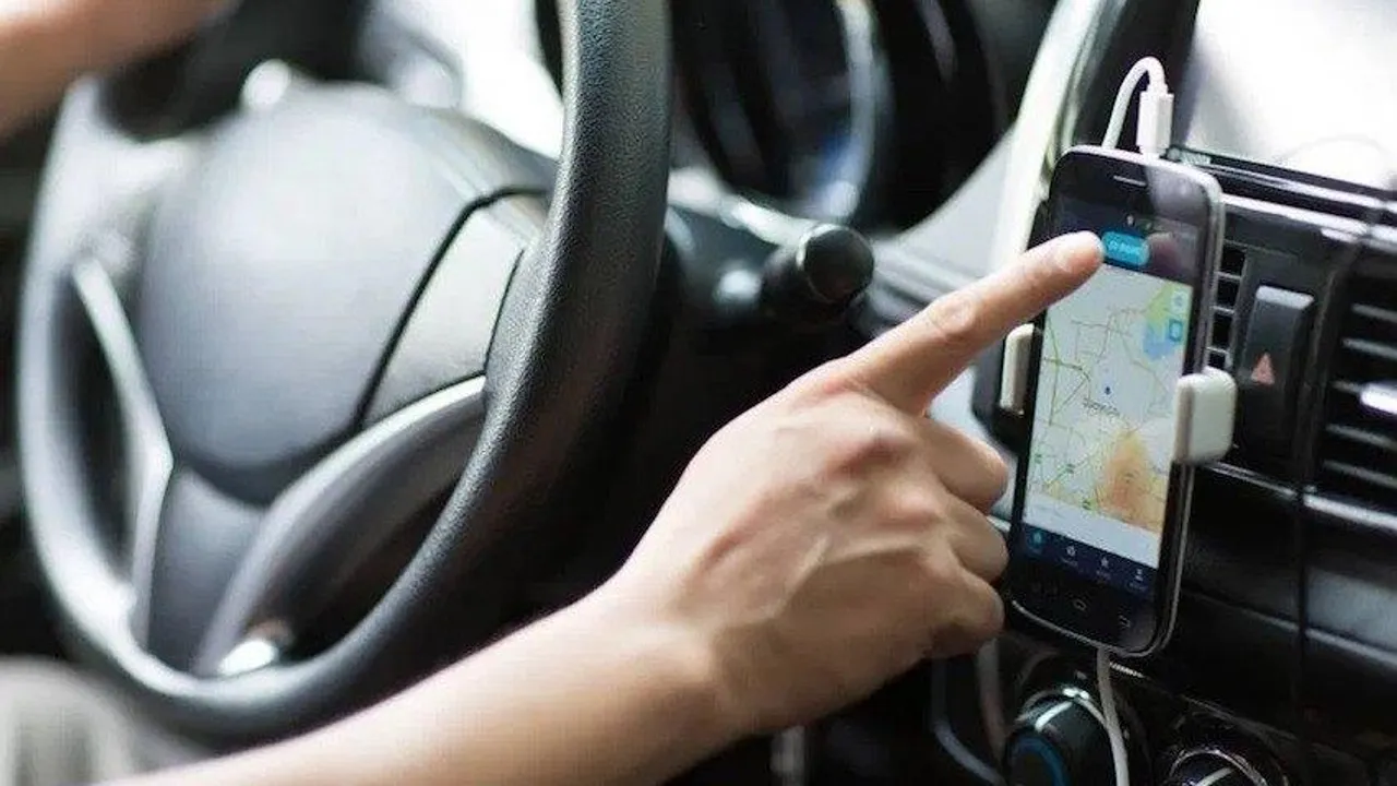 Mano de un chofer al volante manipulando un teléfono móvil con mapa de navegación colocado en el panel del vehículo.
