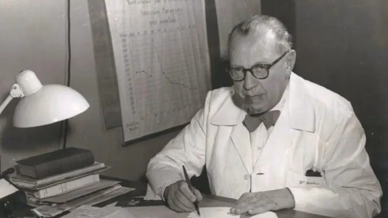 Juan Max Boettner sentado en su escritorio con túnica blanca, anteojos y una pluma frente a documentos y gráficos.