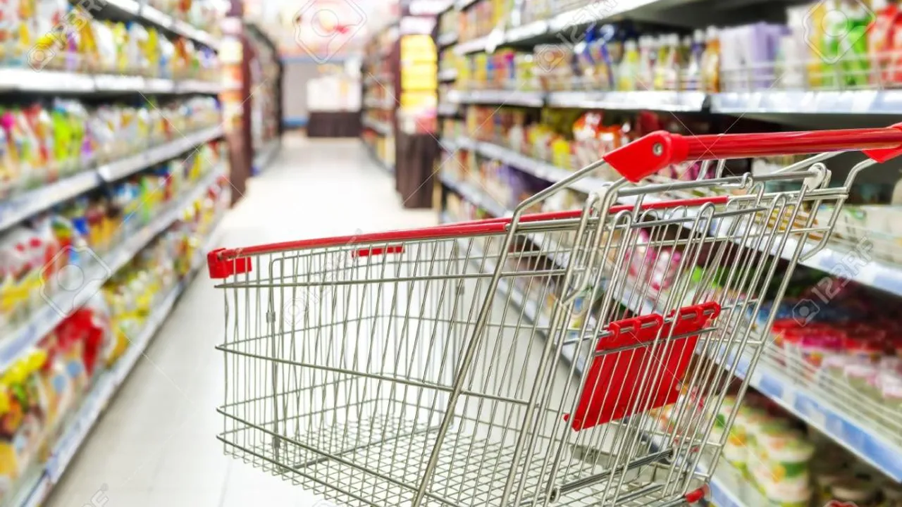 Carrito de compras metálico con manubrio rojo en un pasillo de supermercado con góndolas llenas de productos.