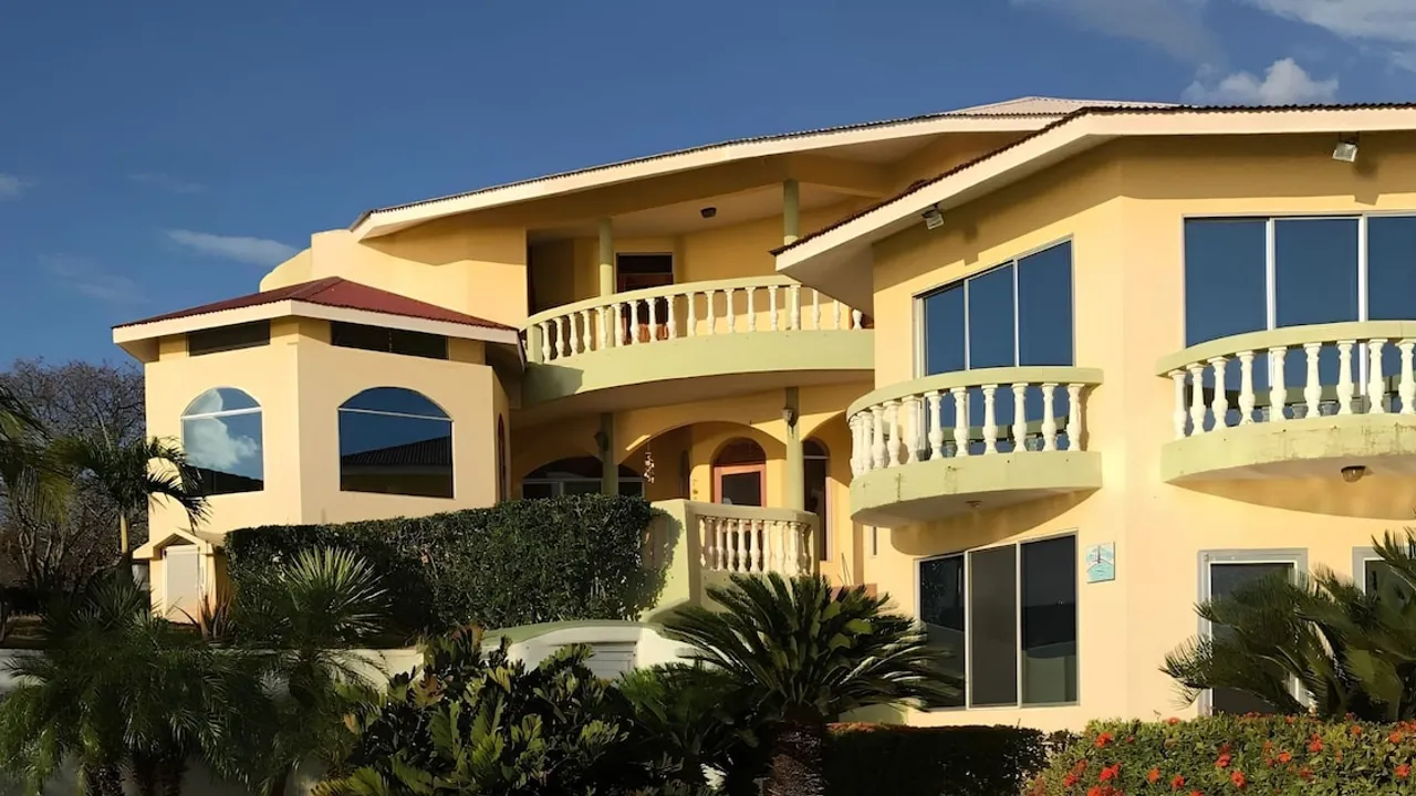 Vivienda de estilo mediterráneo con fachada amarilla, balcones curvos y palmeras bajo un cielo despejado.