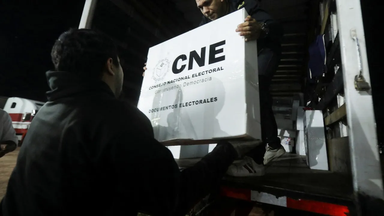 Dos personas cargan una caja blanca con el rótulo del CNE de documentos electorales en un vehículo.