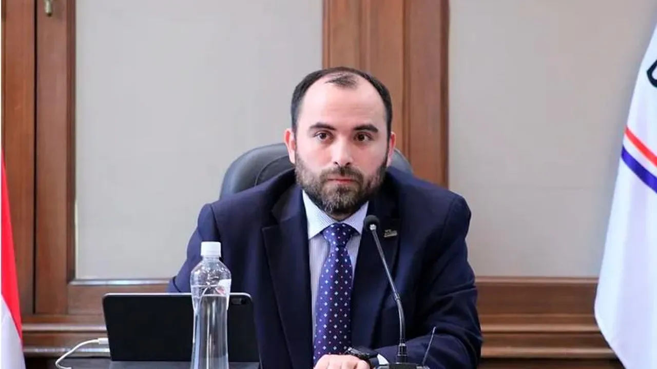 Jorge Bogarín Alfonso con traje oscuro y corbata durante un acto oficial frente a un micrófono y un termo de agua.