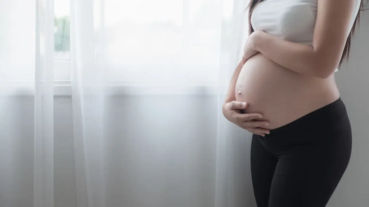 Mujer de perfil en avanzado estado de gestación sosteniendo su vientre descubierto junto a una ventana.