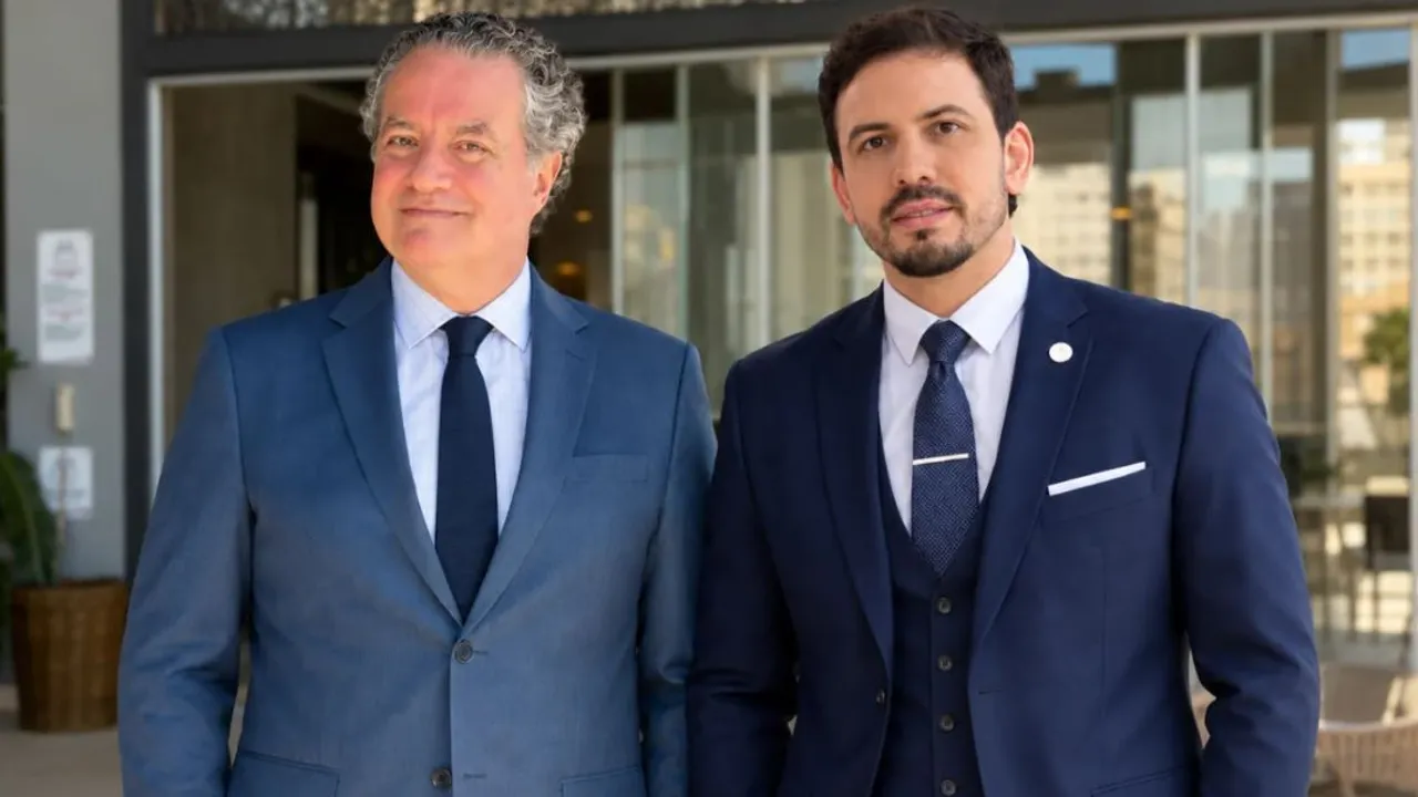 Dos hombres en traje formal de pie al aire libre frente a una entrada vidriada de un edificio.