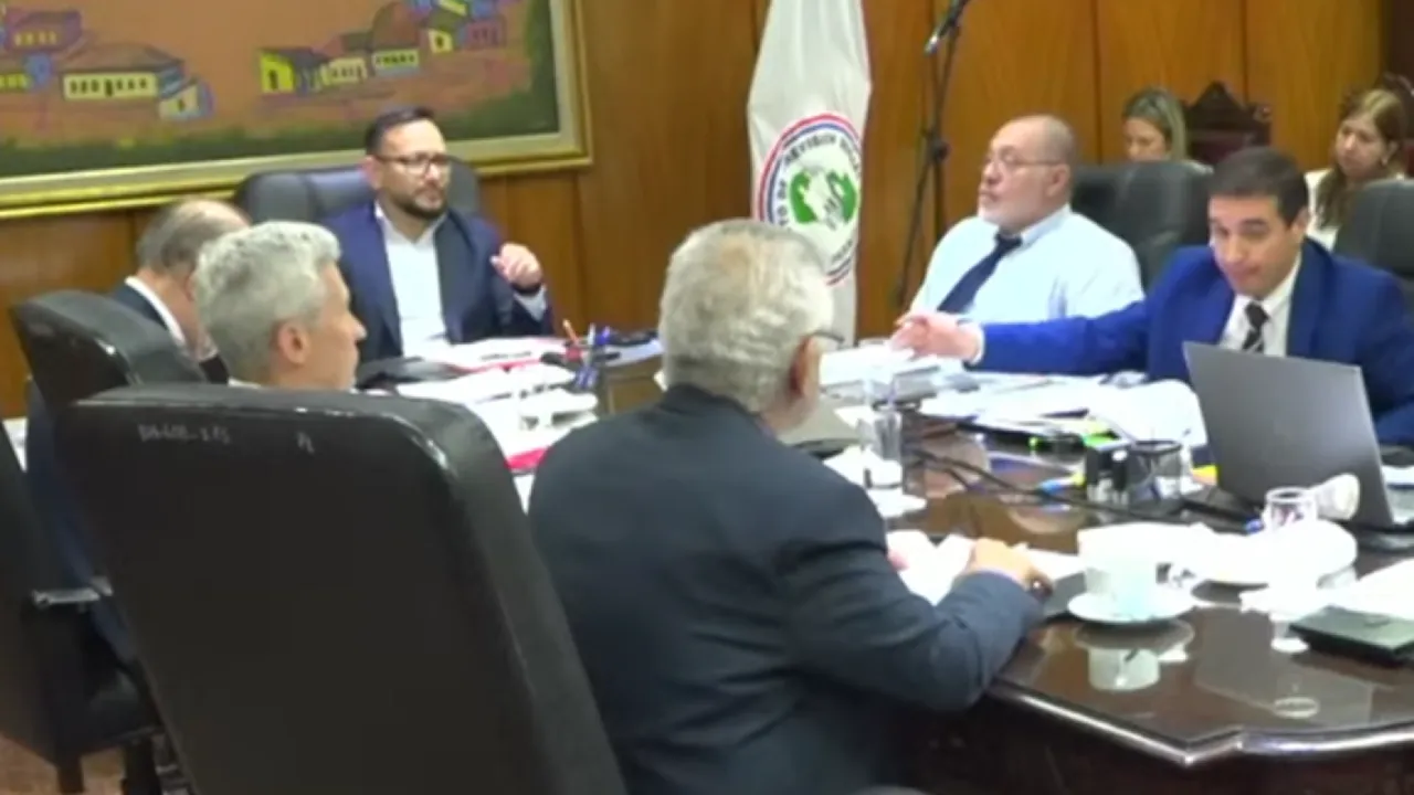 Reunión de autoridades sentadas alrededor de una mesa de directorio con expedientes, una laptop y banderas oficiales.