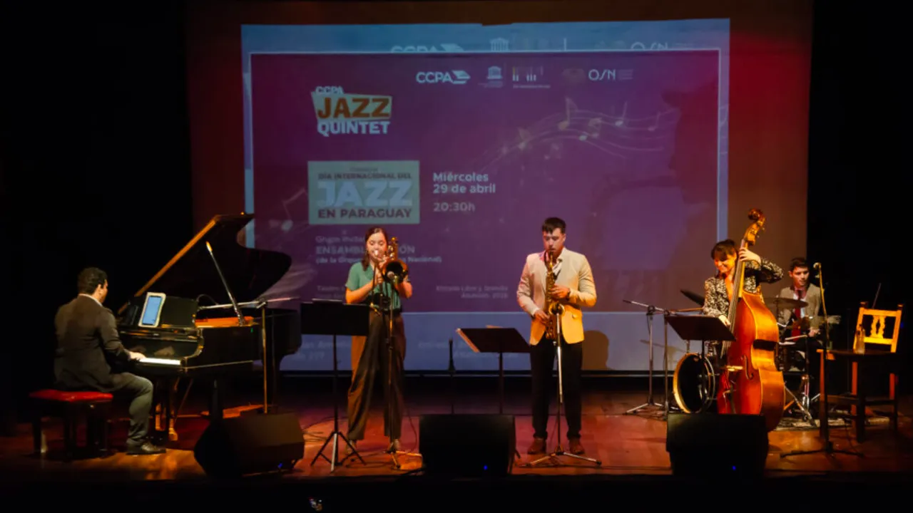 Músicos interpretan jazz en un escenario con piano, saxofón, trombón y contrabajo frente a una pantalla institucional.