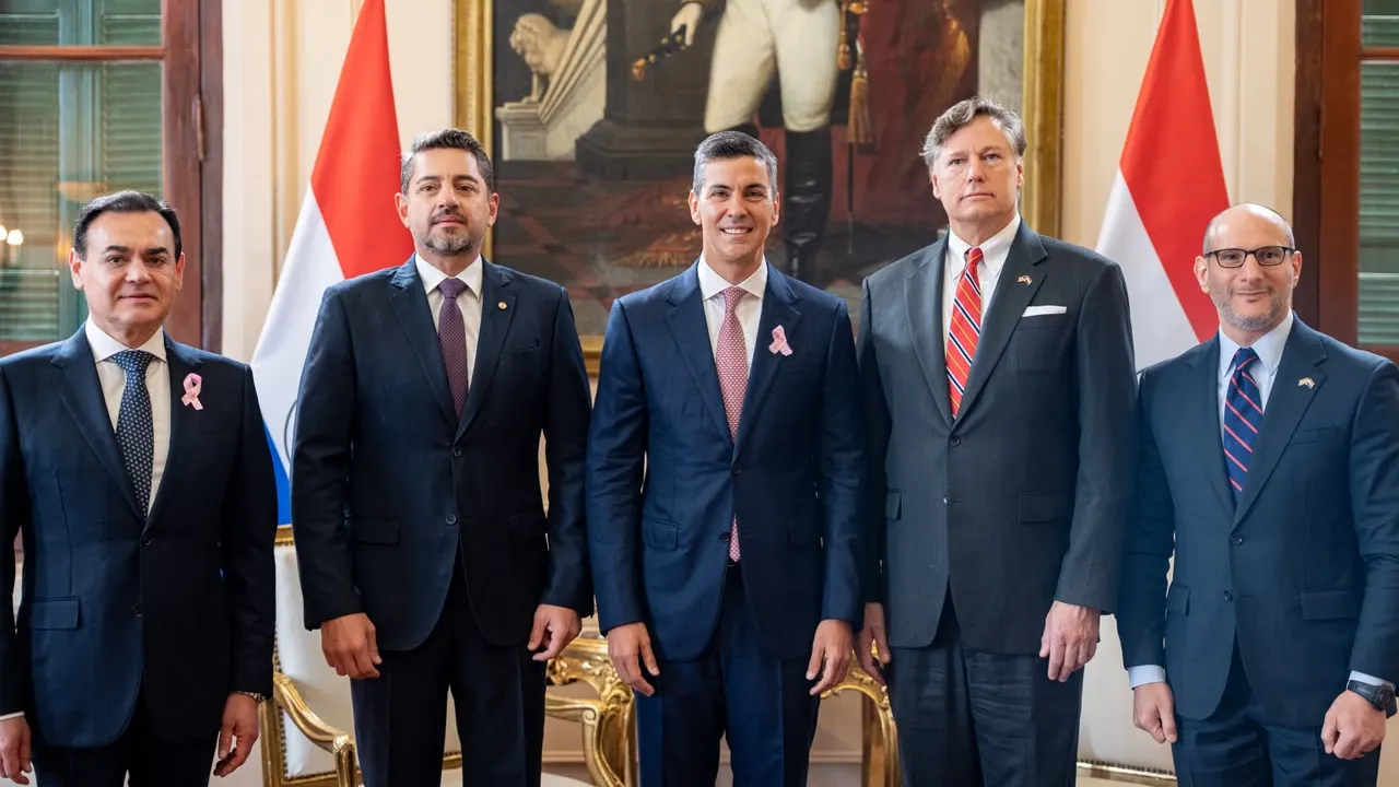 Cinco hombres de traje y corbata posan alineados frente a banderas y un cuadro en un salón oficial.