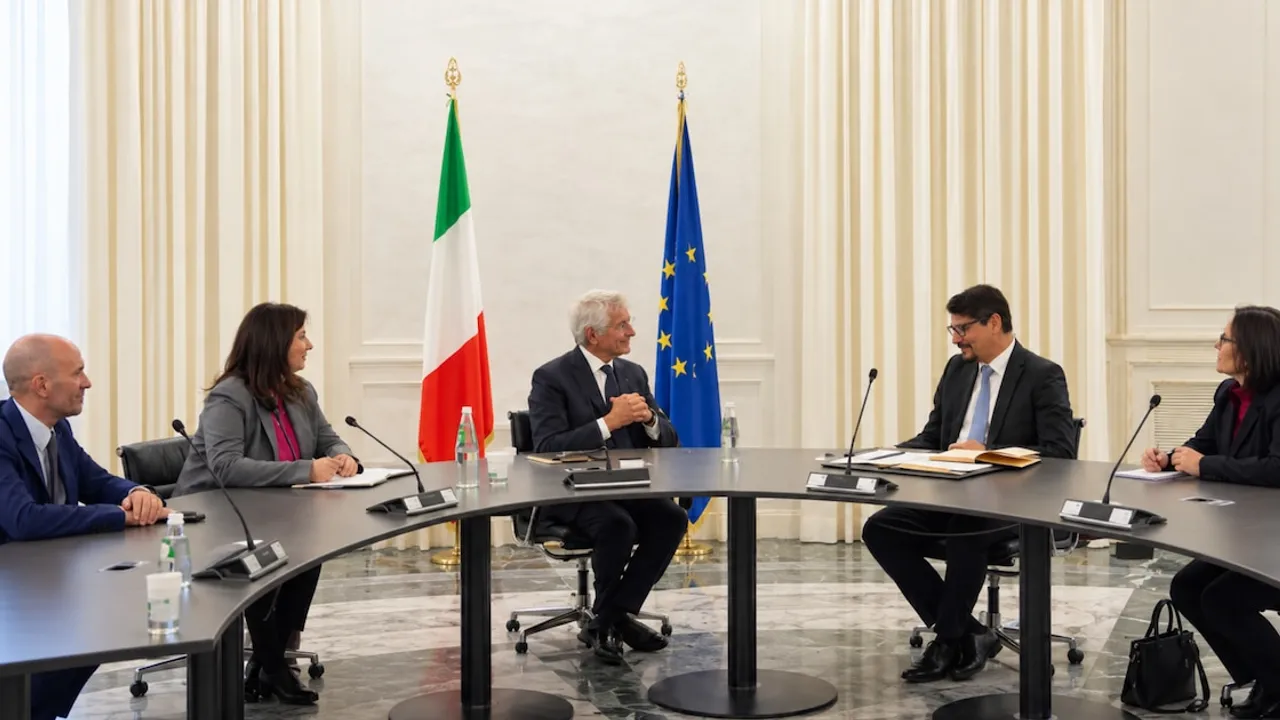 Delegados oficiales conversan alrededor de una mesa negra frente a las banderas de Italia y la Unión Europea.