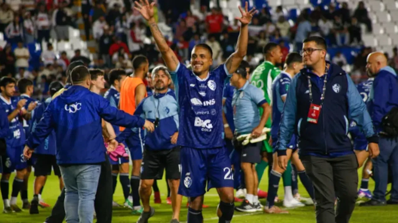 Futbolista con uniforme azul celebra con los brazos en alto rodeado por compañeros en la cancha