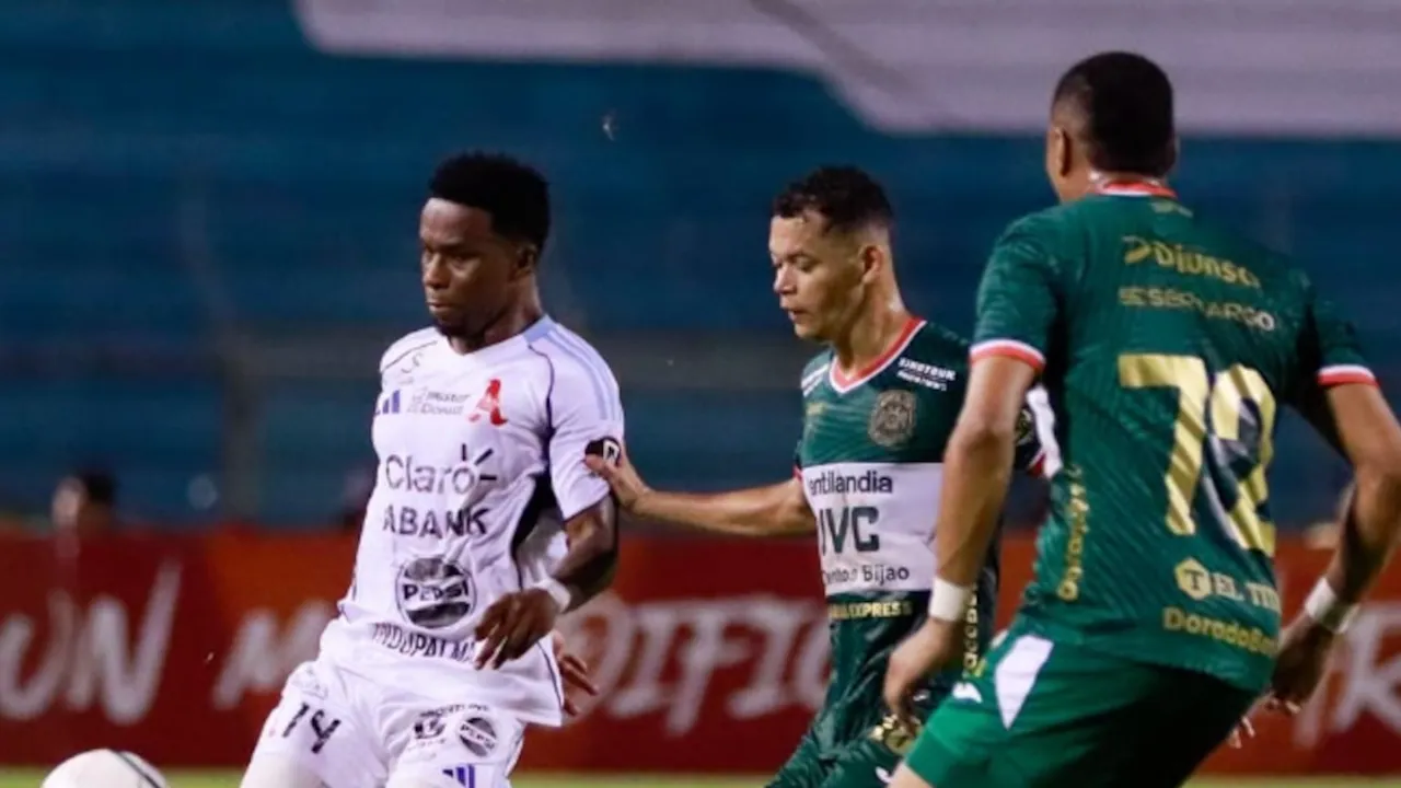 Futbolista con camiseta blanca domina el balón ante la marca de dos rivales con uniforme verde en el campo.