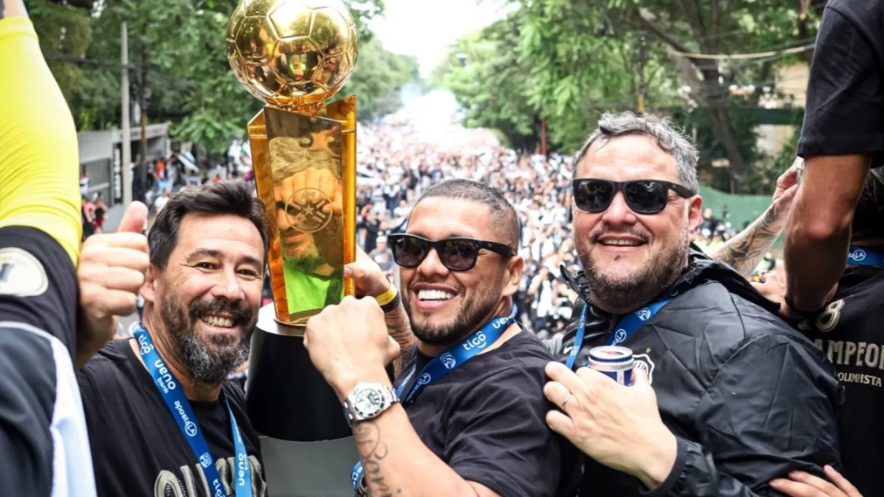 Personas sonrientes sostienen en alto un trofeo dorado de campeón durante una caravana festiva.