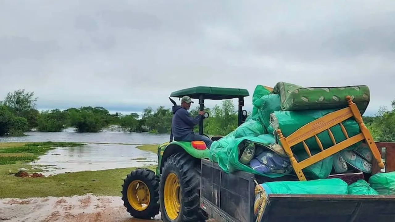 Un hombre conduce un tractor verde cargado con camas y pertenencias frente a un campo anegado por el agua.