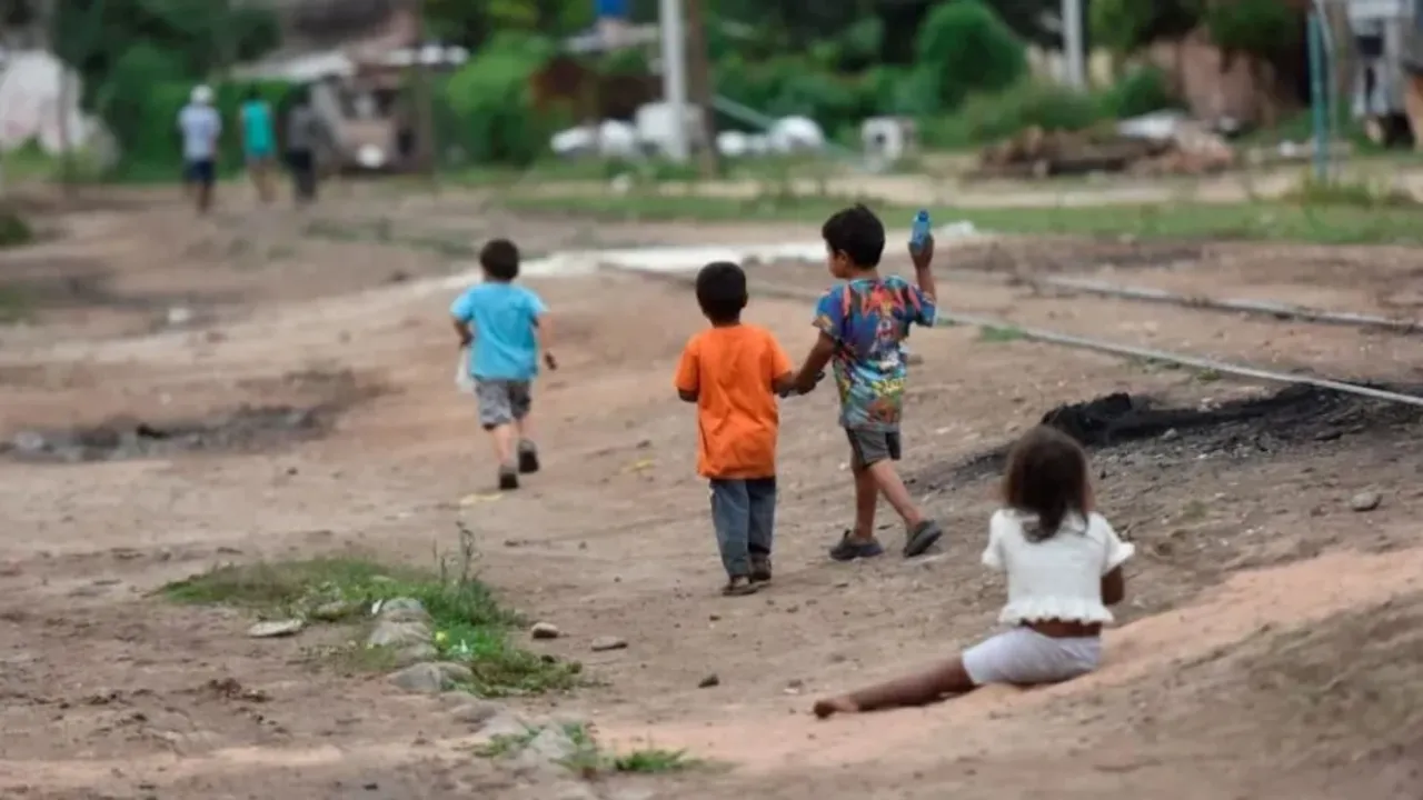 Varios niños caminan y juegan descalzos por una calle de tierra cerca de unas vías de tren.