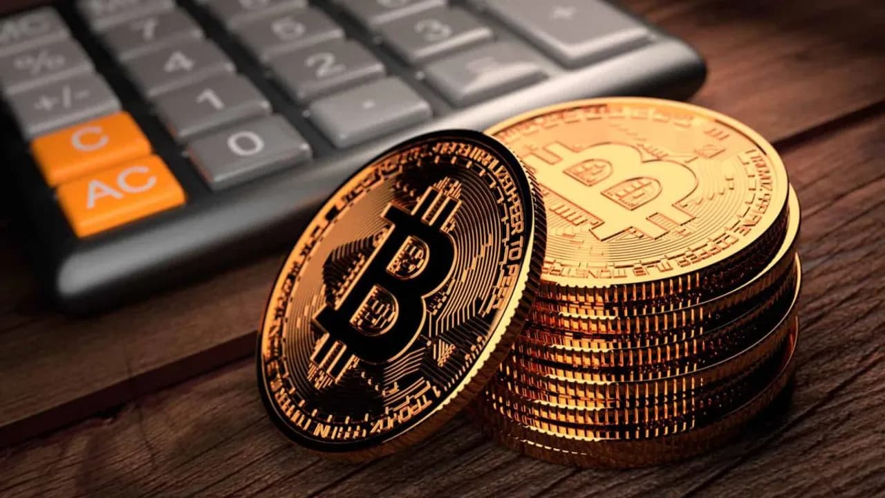 Monedas doradas de bitcoin apiladas junto a una calculadora sobre una mesa de madera oscura.