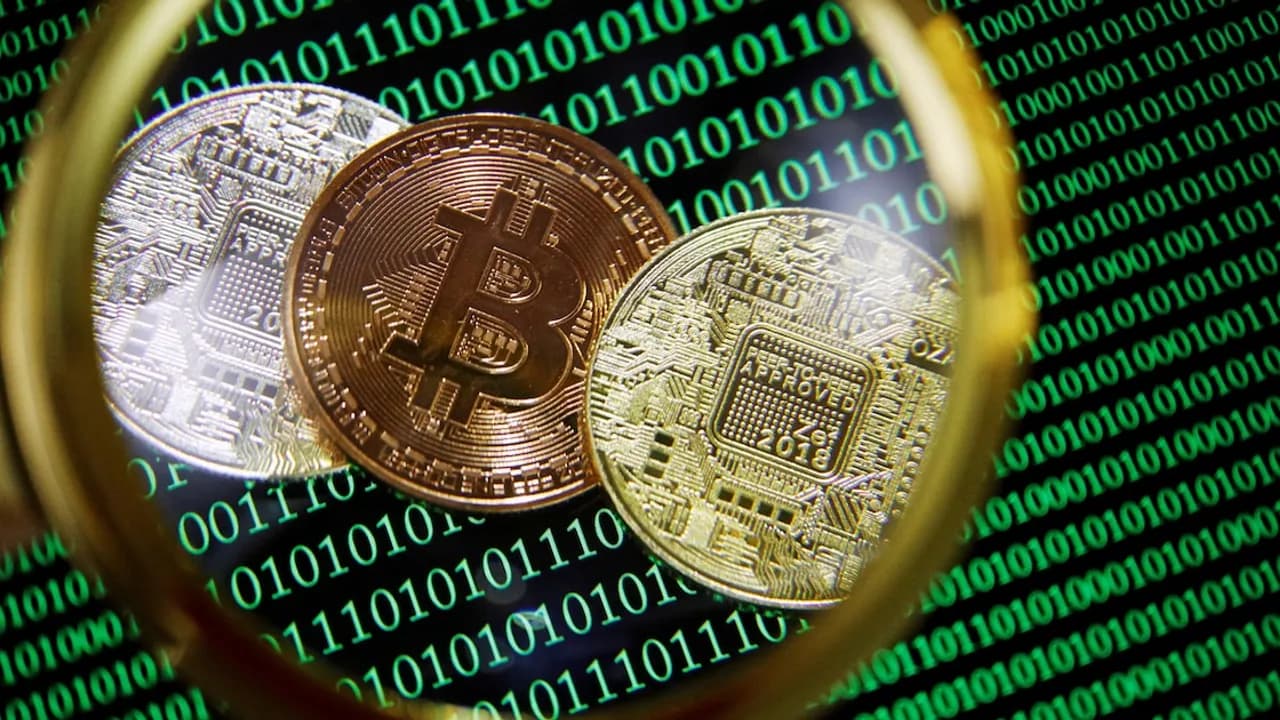 Monedas físicas de bitcoin sobre una pantalla con código binario verde vistas a través de una lupa.