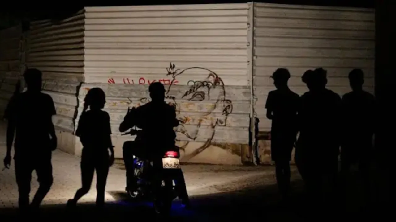 Siluetas de varias personas y una motocicleta en una calle a oscuras frente a una pared de chapa con grafitis.