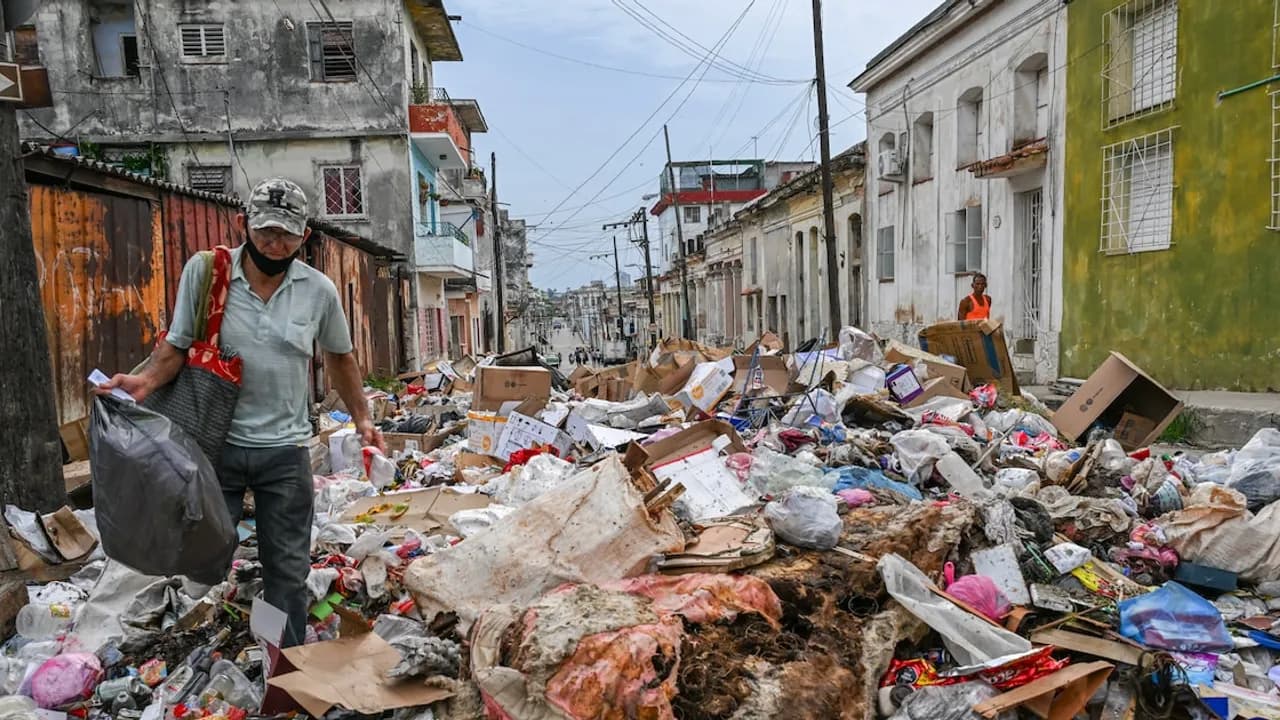 Hombre camina junto a montículos de basura acumulada y cajas de cartón en una calle deteriorada con edificios antiguos.