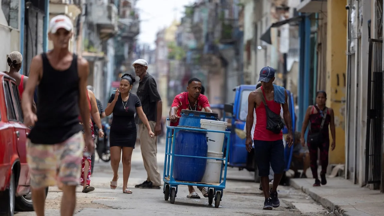 Personas caminan por una calle precaria mientras un hombre empuja un carro con bidones plásticos de agua.