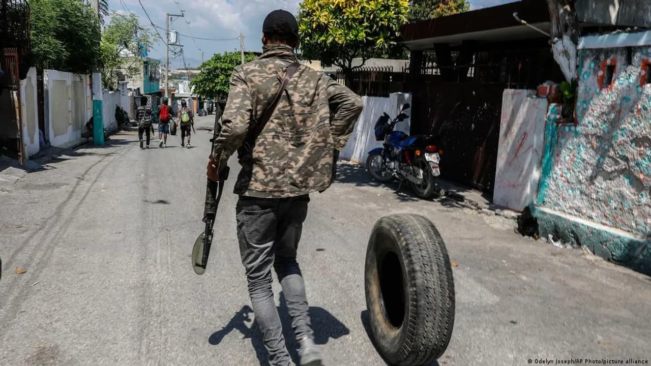 Hombre armado viste ropa de camuflaje y camina por una calle empedrada empujando un neumático usado.