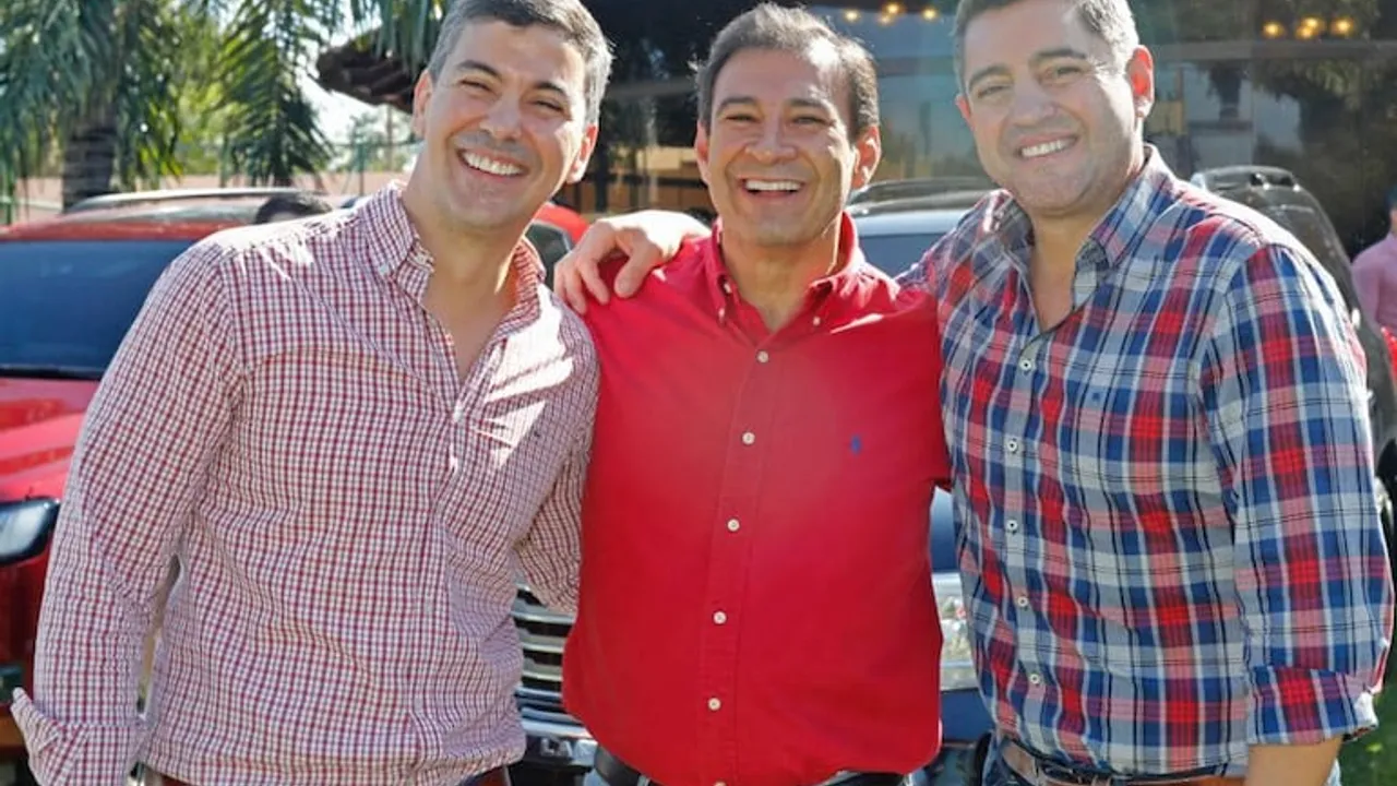 Santiago Peña, Silvio Ovelar y Pedro Alliana sonríen abrazados al aire libre frente a varios vehículos estacionados.