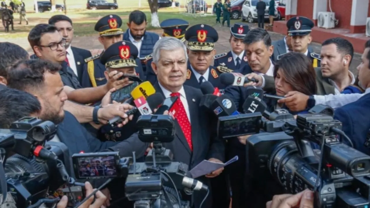 Un hombre con traje y corbata roja habla frente a numerosos micrófonos y cámaras de prensa rodeado de uniformados.