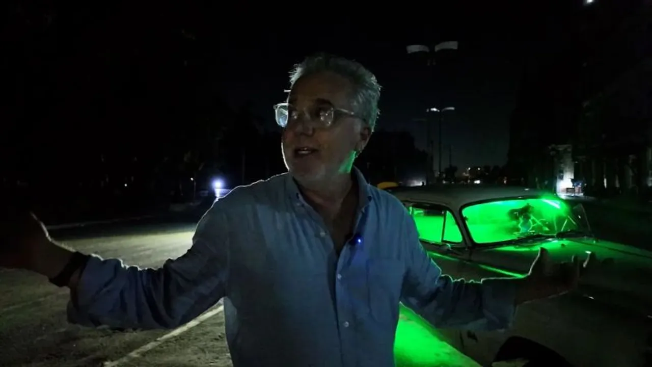Un hombre con anteojos gesticula de noche frente a un auto antiguo iluminado con luces de neón verdes.