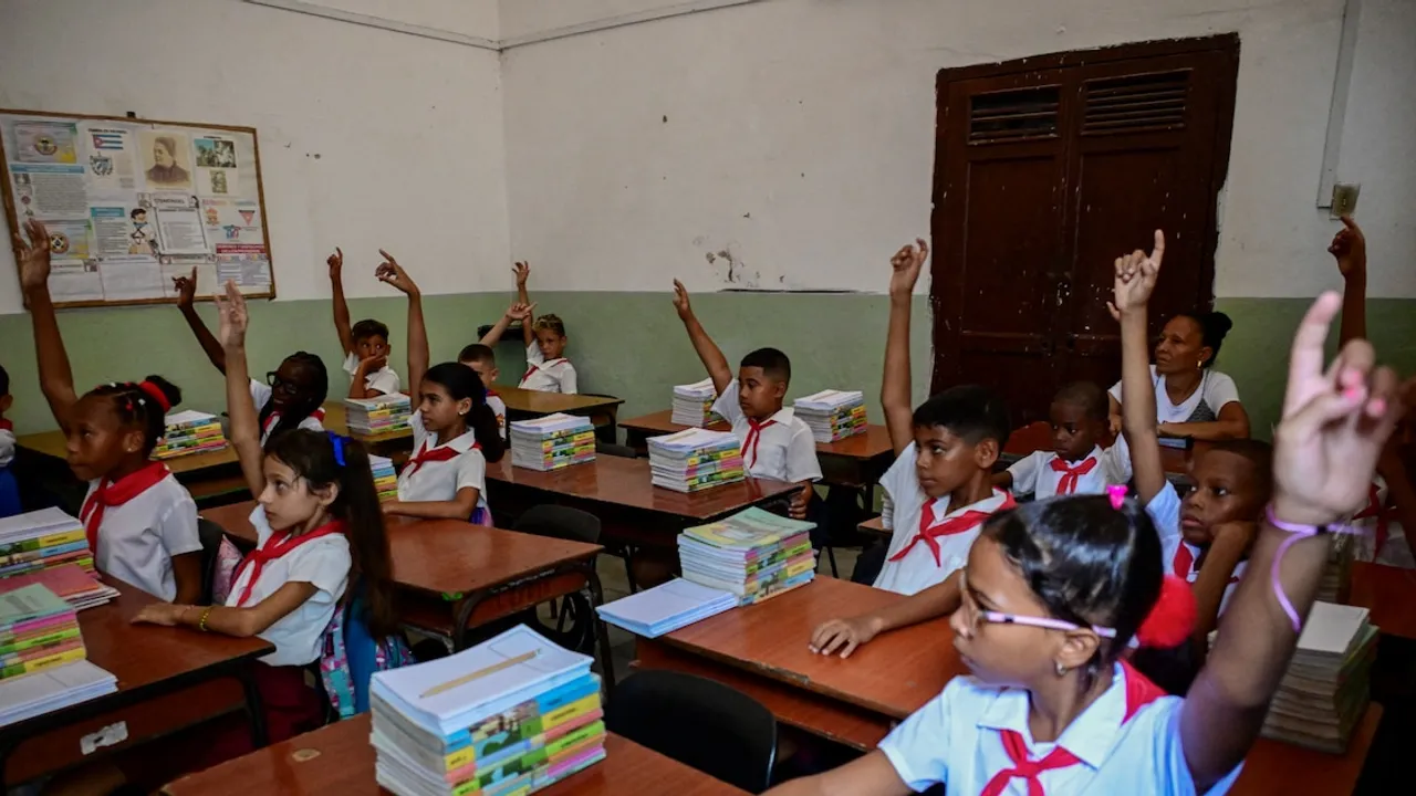 Estudiantes en un aula escolar sentados en pupitres con libros levantan la mano durante una clase.
