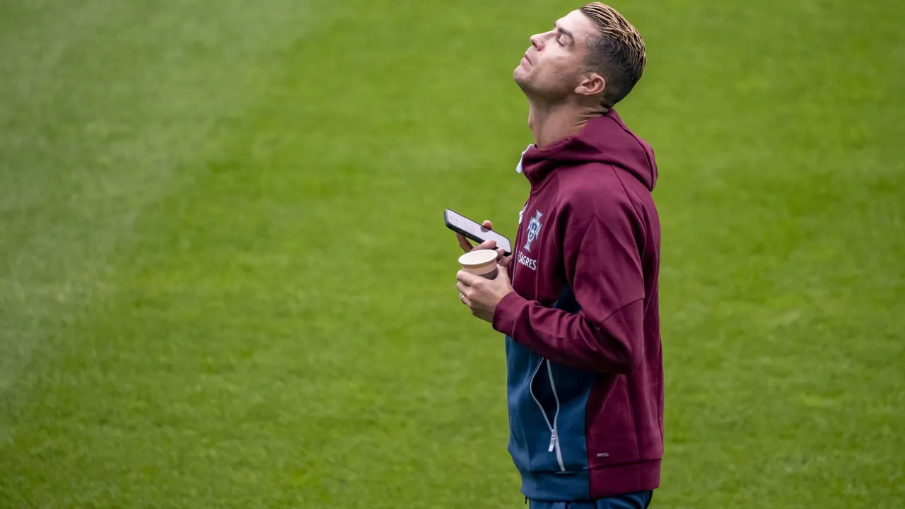 Cristiano Ronaldo con sudadera granate de la selección de Portugal mira hacia arriba sosteniendo un teléfono y un vaso.