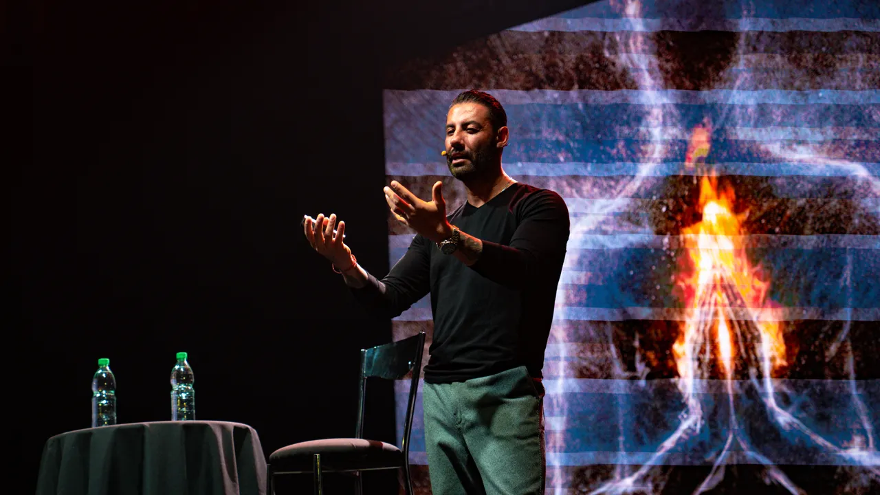 Daniel Dueñas habla sobre un escenario con micrófono de solapa y una pantalla con fuego de fondo.
