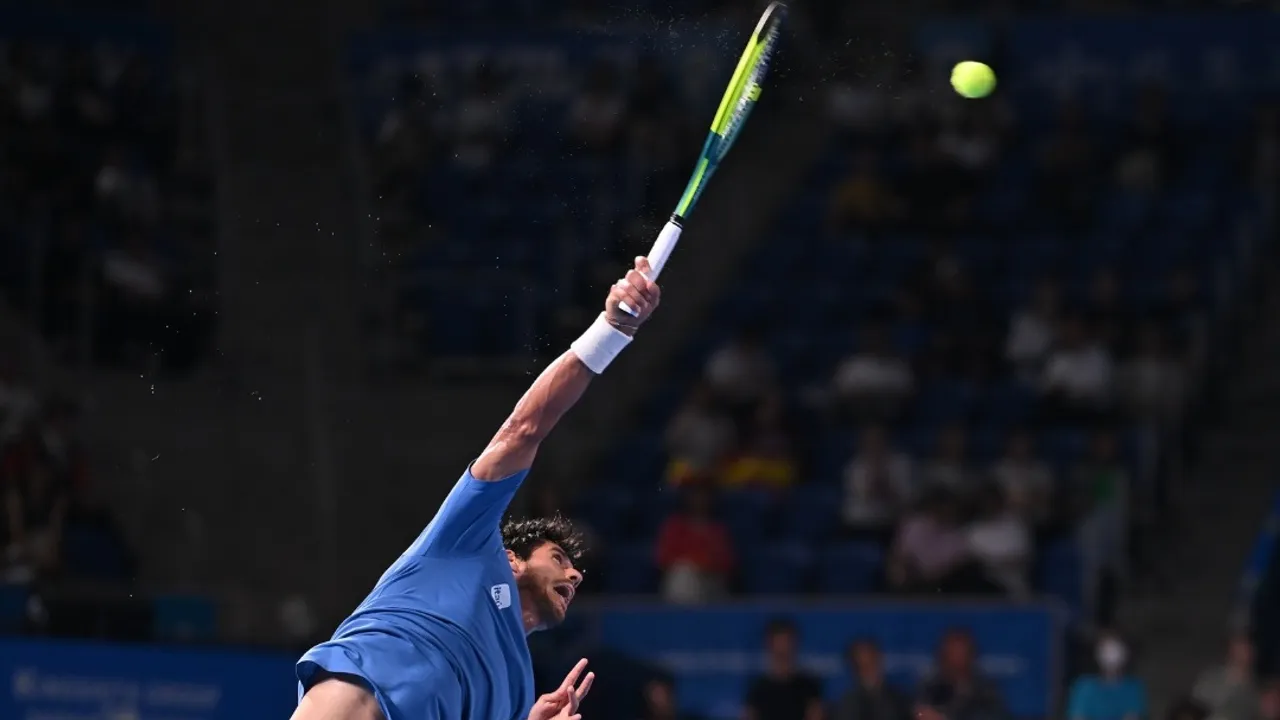 Tenista con camiseta azul estira el brazo para ejecutar un saque de tenis en una pista dura profesional.