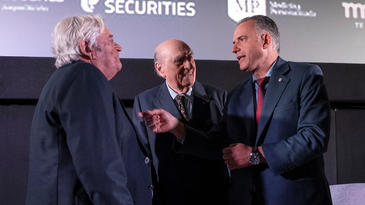 Tres hombres vestidos de traje conversan de pie en un evento institucional frente a una pantalla de fondo.