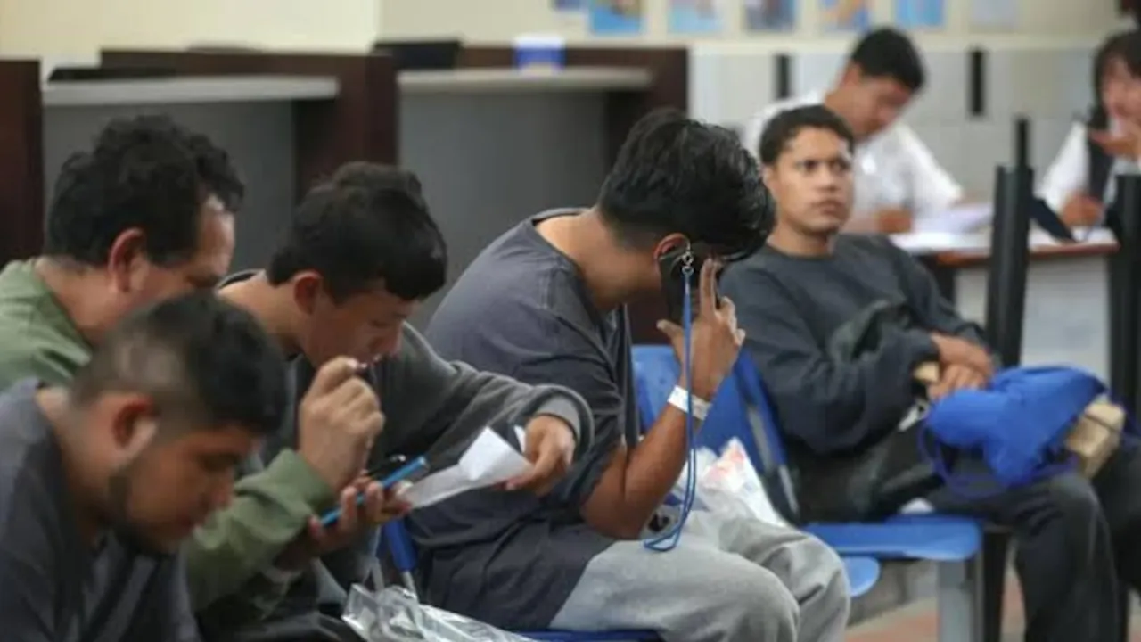Varios hombres sentados en una sala de espera con documentos y teléfonos móviles durante un trámite migratorio.