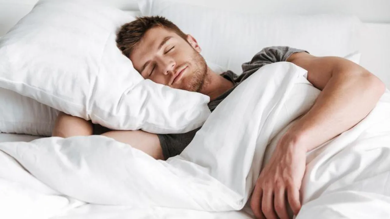 Hombre acostado en una cama descansando con la cabeza sobre una almohada blanca y tapado con una sábana clara.
