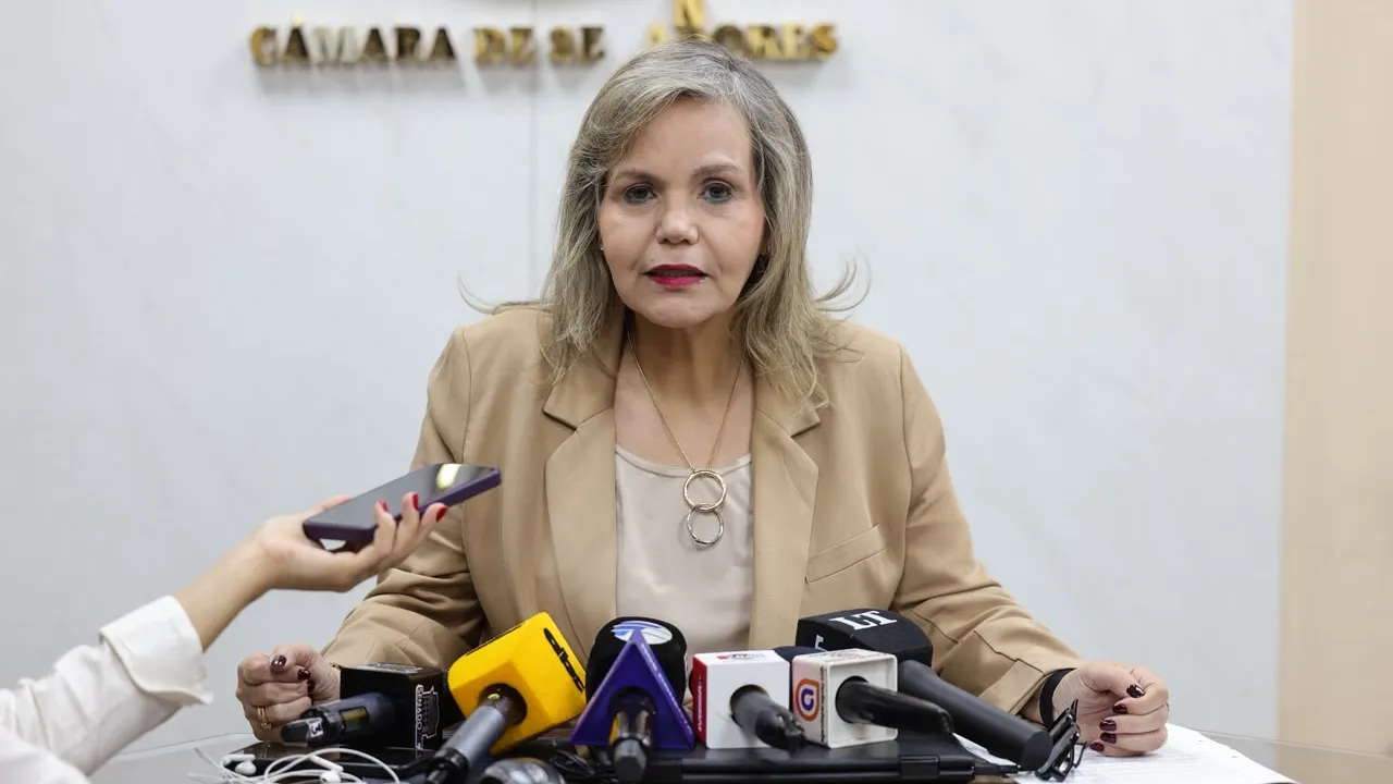 Mujer rubia con saco beige sentada ante micrófonos de prensa en una sala de conferencias institucional