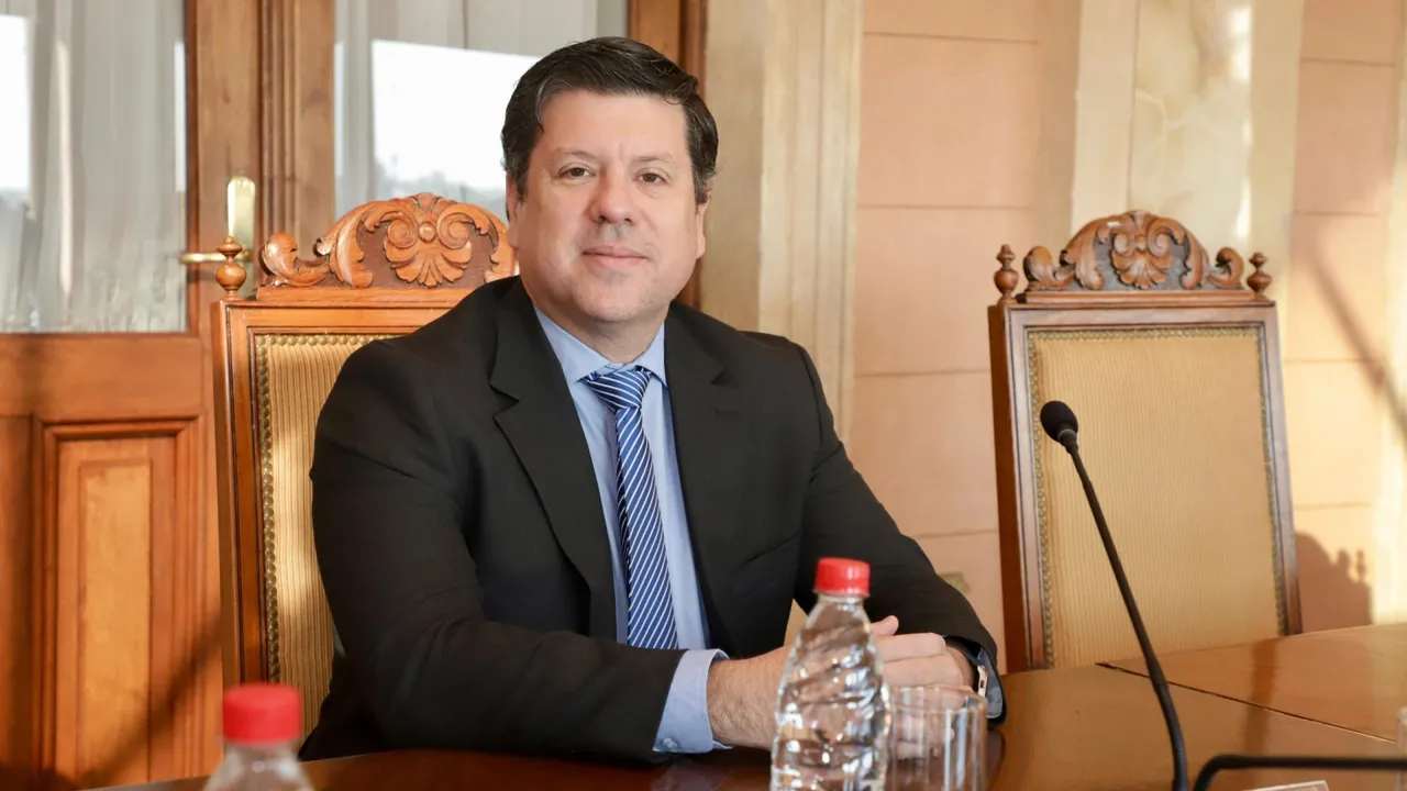 Javier Giménez con traje oscuro y corbata a rayas sentado en una mesa de reuniones con micrófonos y botellas de agua.