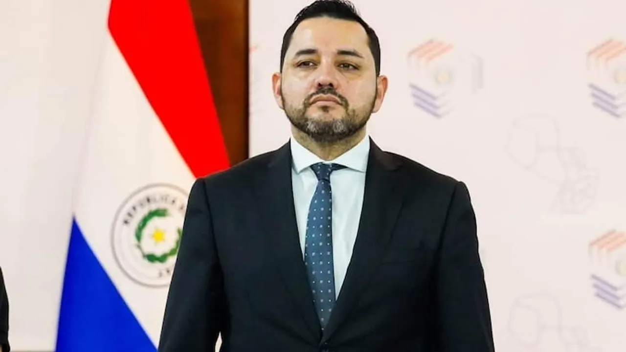 El intendente Luis Bello viste traje oscuro y corbata con la bandera de Paraguay de fondo en un acto oficial.