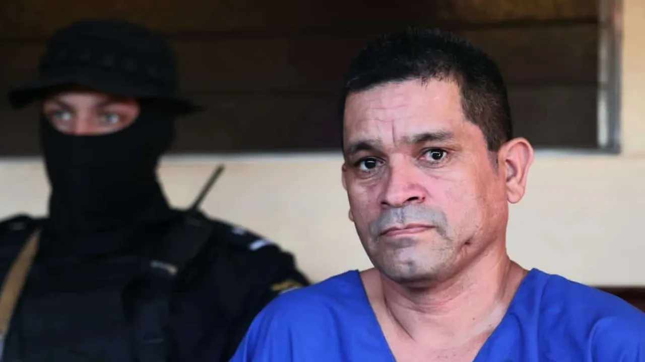 Hombre de remera azul custodiado por un uniformado con pasamontañas negro en una sala.